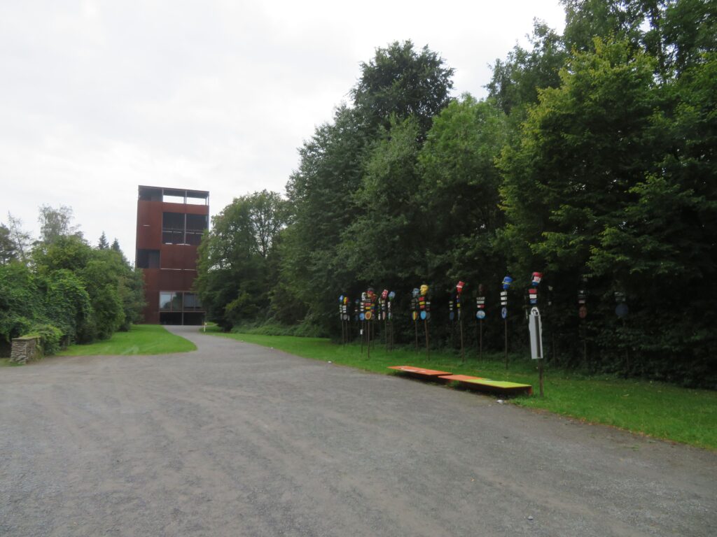 Ein dunkelroter Turm steht am Ende des Weges. Der Turm ist rechteckig und hat große Fenster. Rechts sind viele Bäume und davor stehen Stangen mit Masken. Die Masken haben die Farben verschiedener Länder.