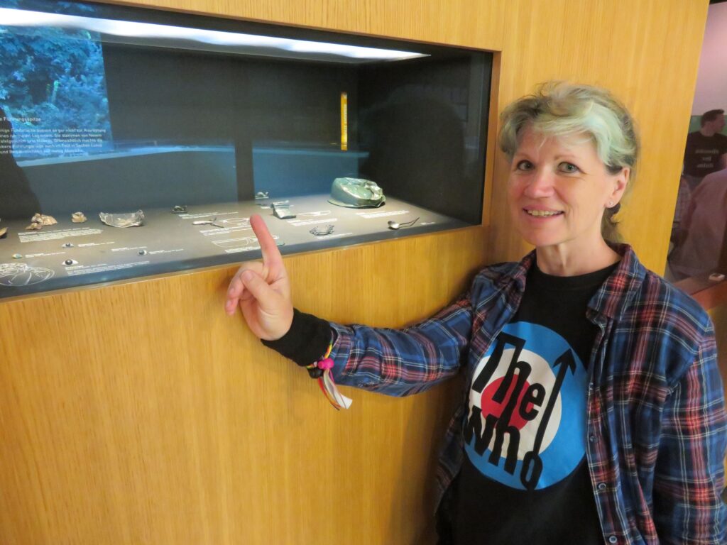 Sandra steht mit einem The Who Shirt und einem karrierten Hemd vor einer Vitrine und zeigt auf eine kleine Delfinfigur, der zusammen mit anderen Ausstellungsstücken hinter Glas liegt.