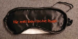 Eine schwarze Schlafmaske auf der mittig in Orange der Text "Für mei bayrische Ruah" gestickt ist.