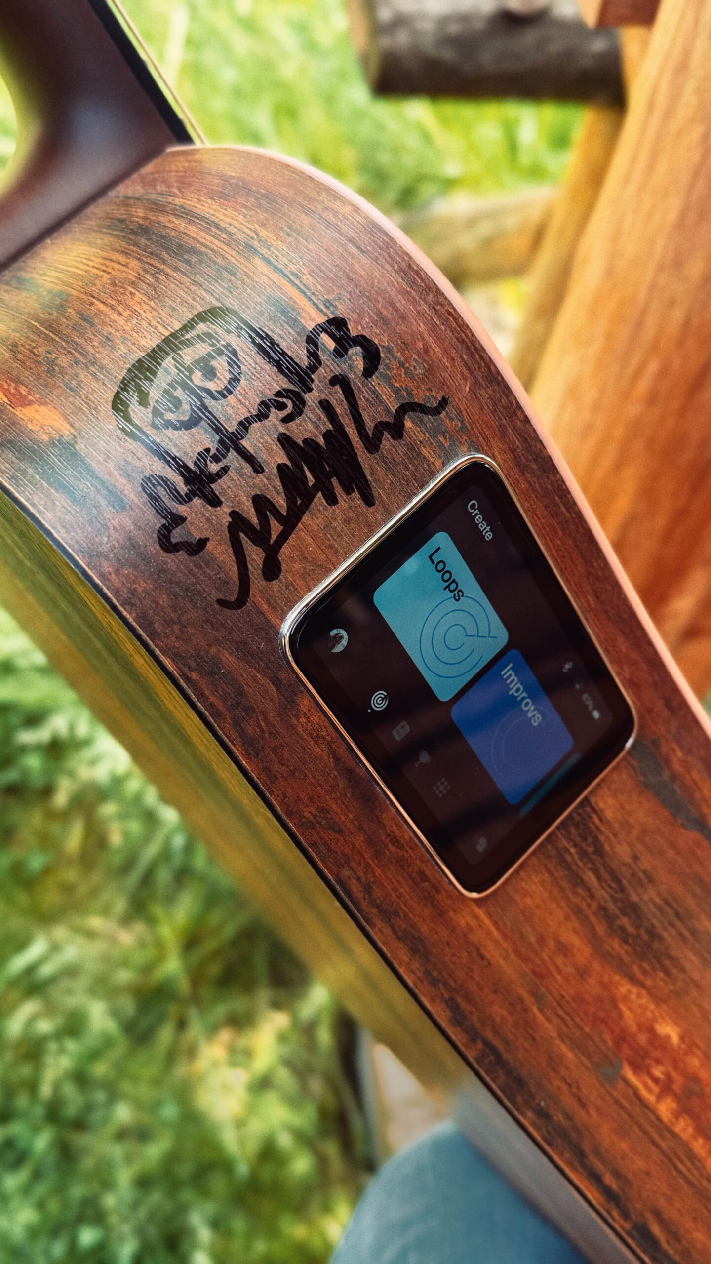 Seitlich am Körper der Gitarre ist ein Autogramm von Tommy mit einem gezeichneten Bernd. Daneben ein in die Gitarre integriertes Touchdisplay, auf der oben "Create" steht. Darunter zwei große Schaltflächen mit "Loops" und "Improvs" und darunter eine Reihe weitere Icons.