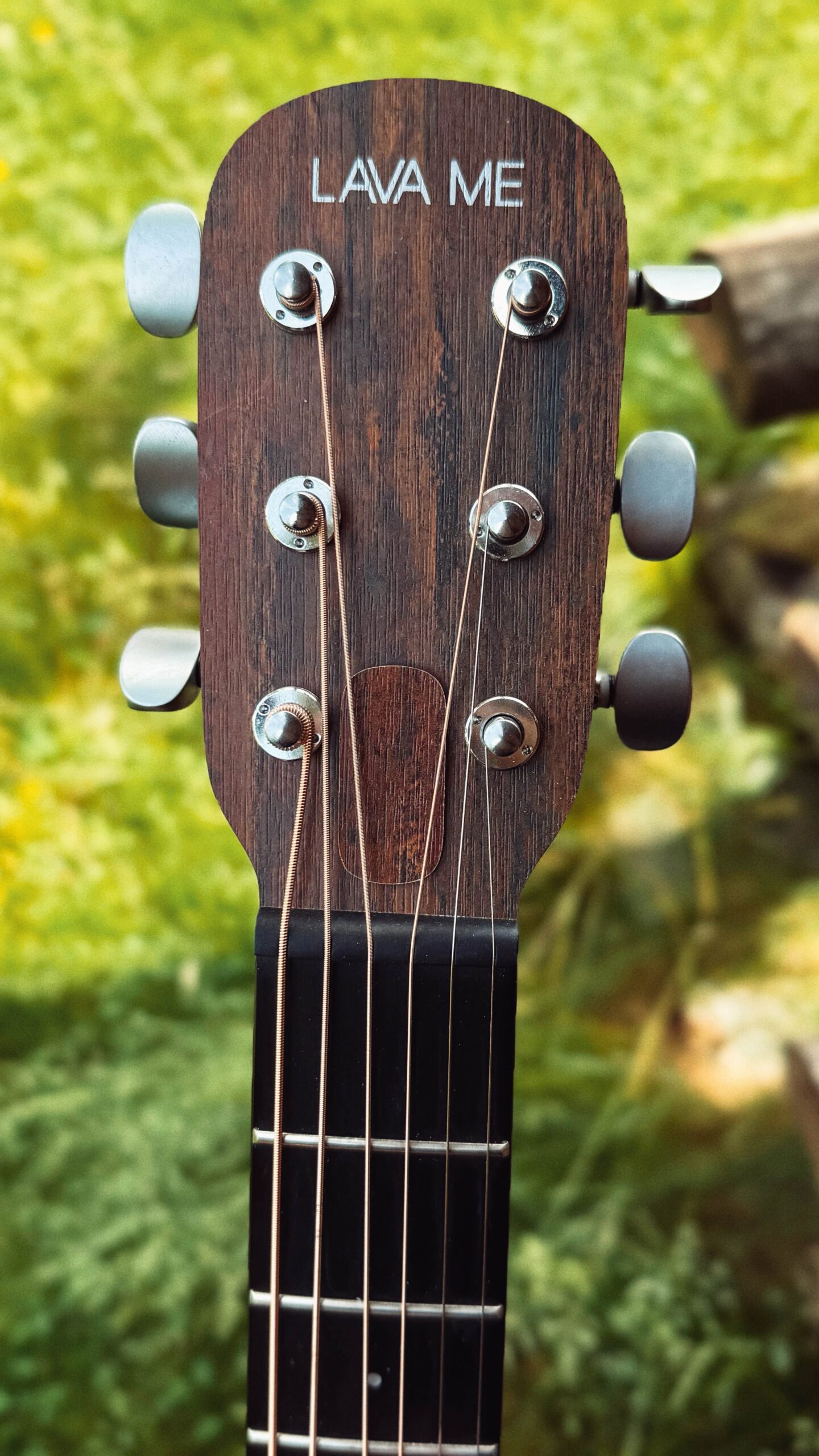 Kopf der Gitarre mit dem Schriftzug LAVA ME. Im Hintergrund eine grüne Wiese.