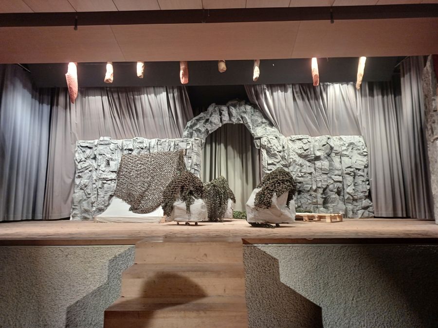 Auf der Bühne steht im Hintergrund eine aus Styropor gebaute Steinwand mit einem Durchgang in der Mitte. Davor Felsen aus Pappmaché. Darüber liegen grüne Tarnnetze. Vor der Bühne ist eine kleine Treppe. Von der Decke hängen gebastelte Stalaktiten.