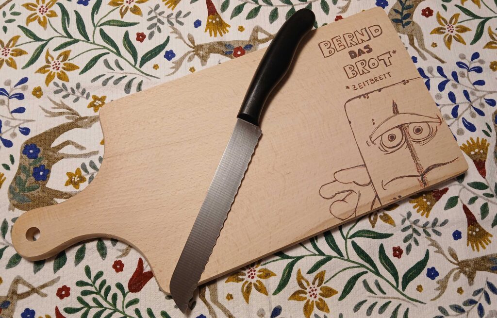 Das Bild zeigt ein Brotzeitbrett aus Holz, auf dem ein gezacktes Brotmesser diagonal liegt – bereit für die nächste Scheibe Käse oder Wurst. In das Brett ist die Figur Bernd das Brot eingraviert: rechteckig, traurig dreinblickend, mit seinem typischen stoischen Gesichtsausdruck. Darüber und darunter steht der Text: „BERND DAS BROT ZEITBRETT“ – eine augenzwinkernde Kombination aus „Brotzeitbrett“ und „Zeitbrett“, die Bernds notorische Abneigung gegen Aktivität und Veränderung humorvoll unterstreicht. Der Hintergrund ist ein bunter Stoff mit Blumen und kleinen Hirschen – fast wie eine Mischung aus Alpenromantik und Kinderzimmernostalgie. Das verspielte Muster kontrastiert wunderbar mit Bernds grummeliger Aura.