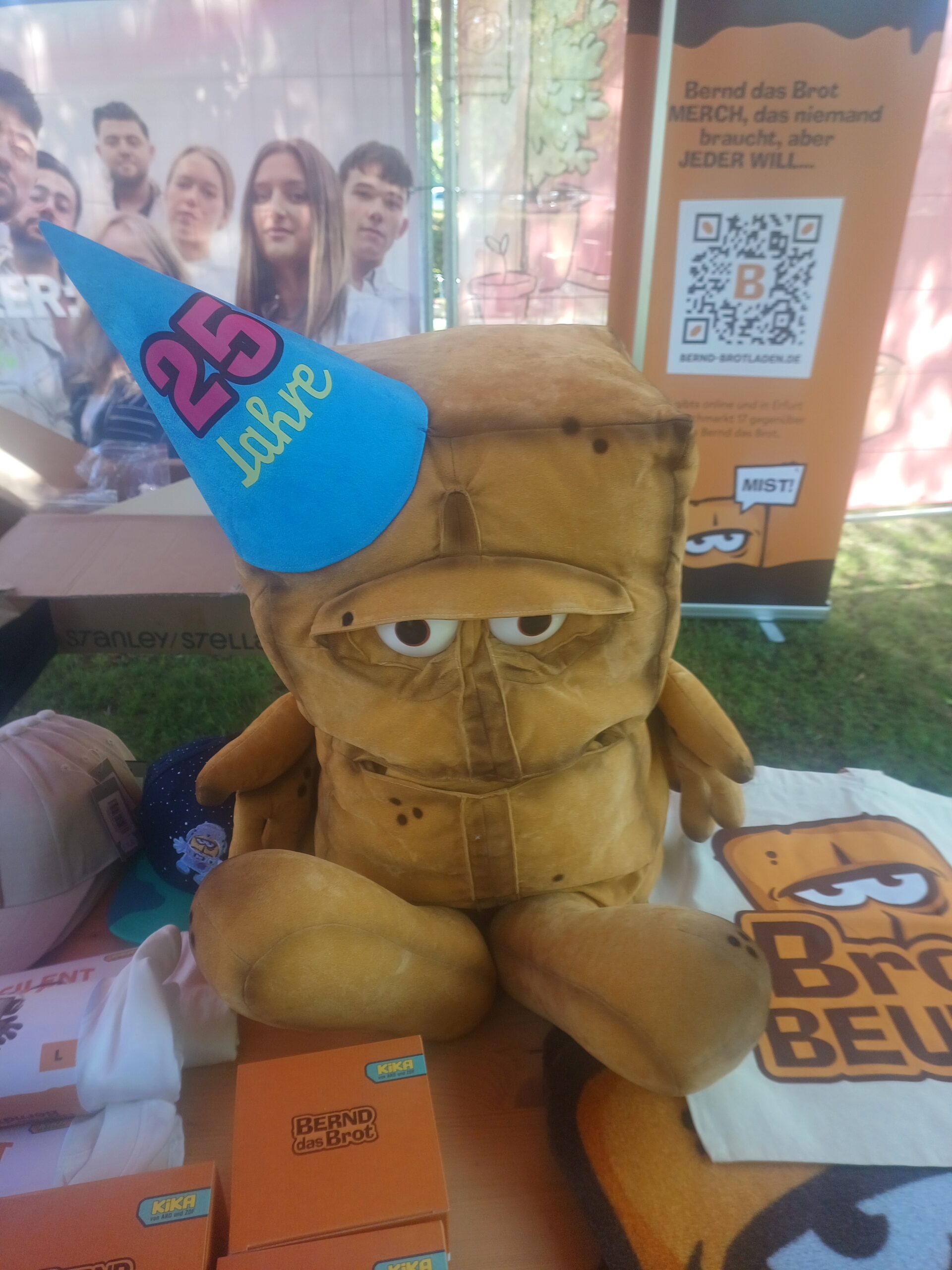 Am Verkaufsstand vom Brotladen sitzt ein 60cm großer Bernd das Brot in der 25 Jahre Jubiläums Edition auf dem Tisch. Neben ihm liegt ein Brotbeutel, stehen Tassen, Shirts und Kappen.