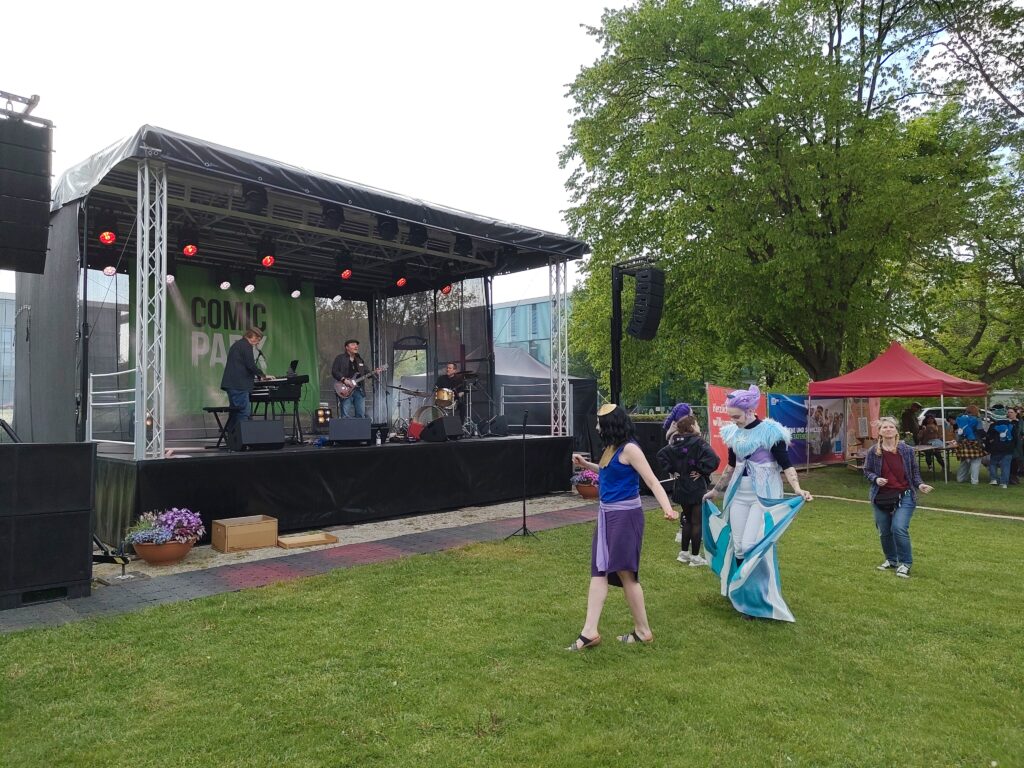 Auf der Bühne ist die Band Harpo Speaks mit Andreas Lenz von Ungern-Sternberg am Keyboard, Tommy Krappweis an der Gitarre und Ingmar Lange am Schlagzeug. Auf der Wiese vor der Bühne tanzen kostümierte Fans.