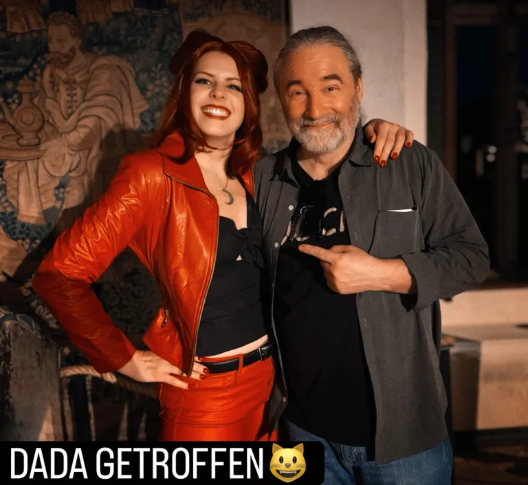 Caro als Dada mit roter Perücke, roter Lederjacke, schwarzem Top mit tiefem Ausschnitt, einer Kette mit einer Mondsichel und schwarzen Lederhosen. Sie lächelt strahlend in die Kamera und hält ihre Hand mit den roten Fingernägeln auf Tommys Schulter, der neben ihr steht, grinst und auf sie zeigt. Unten steht der Text "DADA GETROFFEN" und ein Katzengesichtsmiley. Im Hintergrund ein mittelalterlicher Wandteppich.