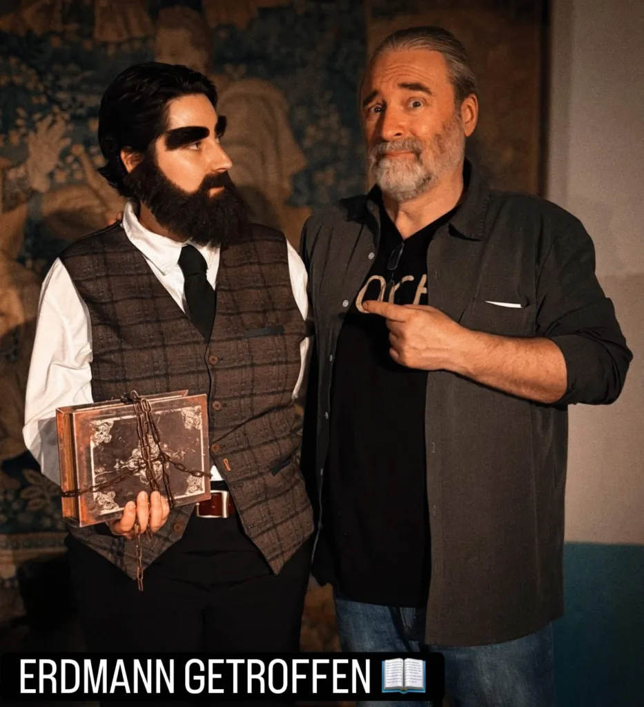 Tommy steht neben Lesensfreude als Erdmann. Sie trägt sehr dichte Augenbrauen und einen künstlichen Vollbart im Gesicht. Zu den schwarzen Hosen und dem weißen Hemd mit schwarzer Krawatte trägt sie eine braun-karierte Weste. In der rechten Hand hält sie ein Buch, das in Ketten gewickelt ist. Tommy zieht die Augenbrauen leicht verzweifelt in die Höhe und zeigt auf Erdmann. Unten steht "ERDMANN GETROFFEN" und das Icon eines Buches. Im Hintergrund sieht man einen mittelalterlichen Wandteppich.