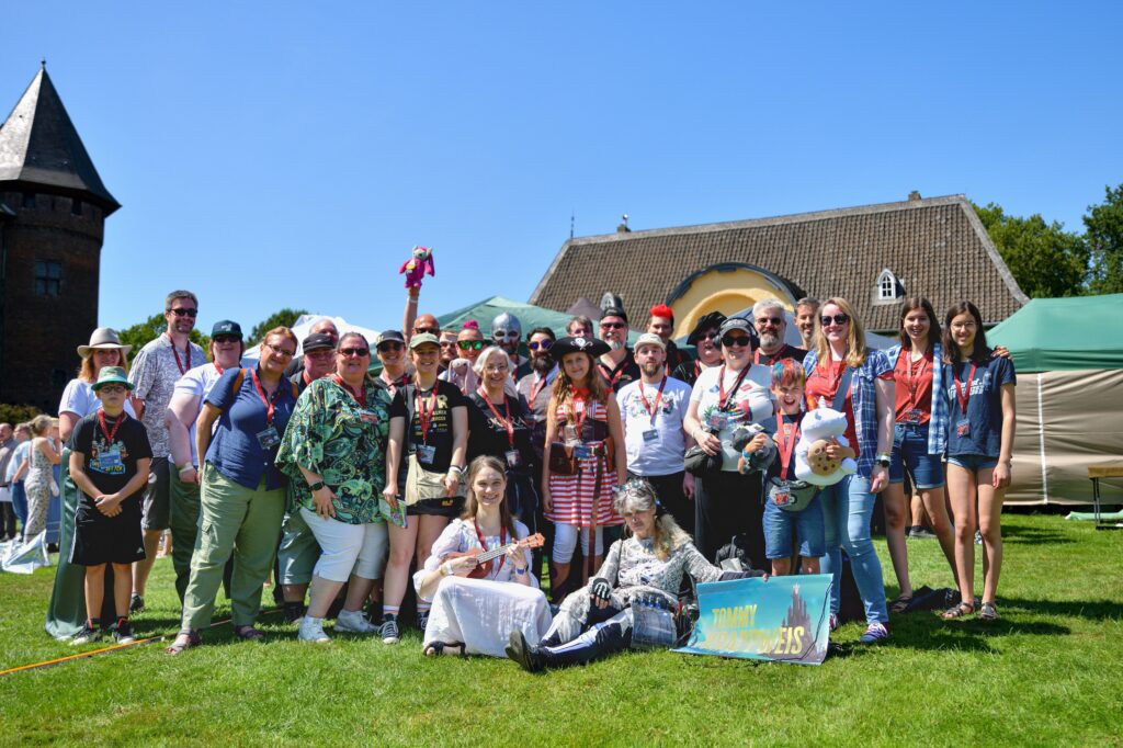 Gruppenfoto von etwa 30 Personen, die teils sommerlich, teils im Cosplay zu sehen sind. Man erkennt mehrere Maras mit rotem Pilzkopf-Shirt, Amalia vom Tannenzweig, ihres Zeichen Oberste Richterin am Europäischen Hexengericht, Spiegeluniversums-Tom mit roten Haaren, Skelett-Hermina Freifrau von Hohenblick frisch aus dem Grab entsprungen, Piratenkapitänin Bonnie McSwan und Geistermädchen Mimi mit Ukulele. Das Mimi-Skelett hält einen Banner mit einem dunklen Berg abgebildet, auf dem der Name TOMMY KRAPPWEIS in gelben Großbuchstaben steht.