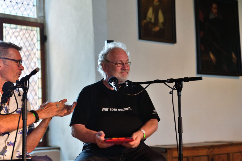 Kai sitzt mit seinem roten iPad auf der Bühne, trägt ein schwarzes T-Shirt mit "War spät gestern." und lächelt über sein Microphonständer hinweg. Links im Bild sieht man Götz klatschen, halb verdeckt durch seinen Microphonständer. Im Hintergrund ist die Burgwand mit kleinkariertvergittertem Fenster, zwei Ölgemälden und einer Holztruhe zu sehen.
