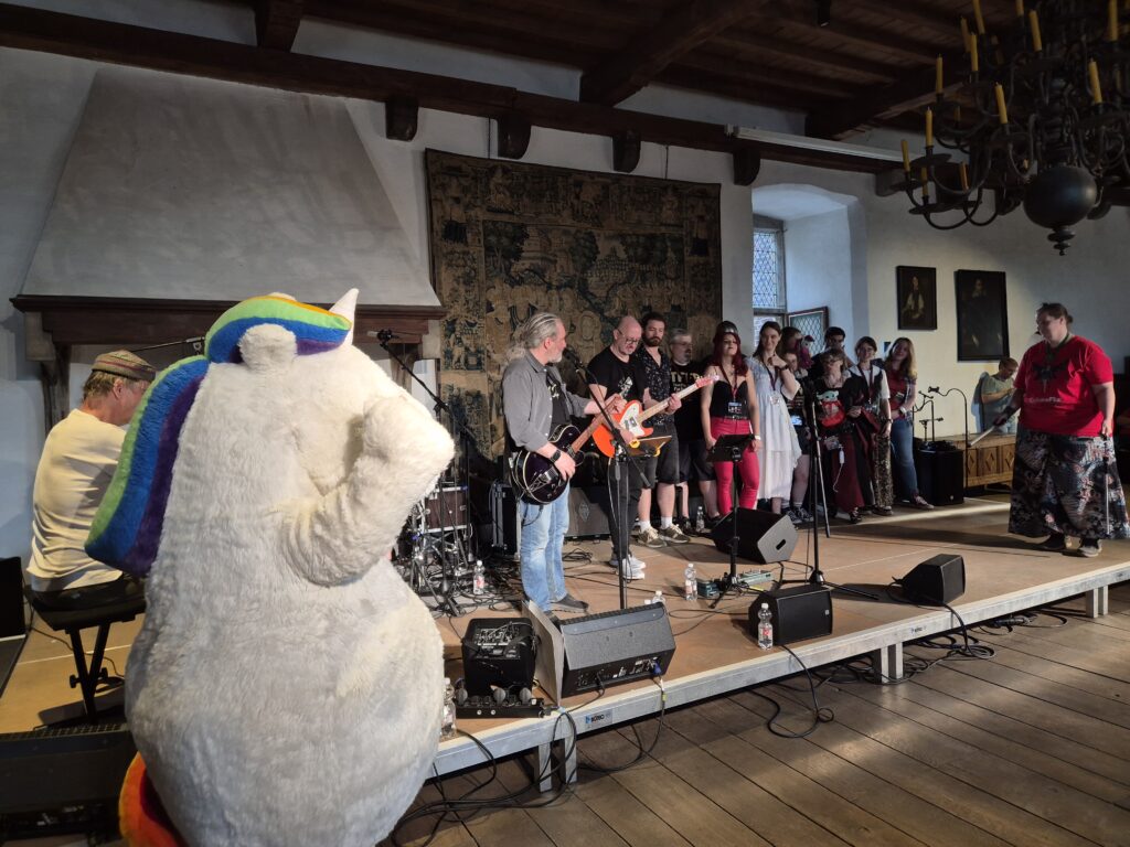 Der Walking Act vom Pummeleinhorn steht for der Bühne, auf der die bumm film Band spielt. Mit auf der Bühne stehen einige Maraverse-Besucher und Heike von der KrähenFee mit LED Poi Sticks. Steffi verteilt u.a. ein Pummeleinhorn-Kissen.