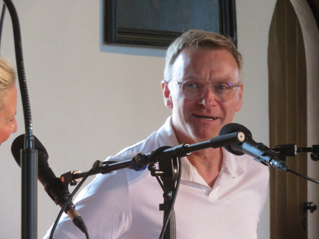 Thomas Nicolai lächelt. Er sitzt mit weißem Poloshirt vor zwei Microphonen. Links sieht man noch die Nase und die lächelnden Augen von Arlett ins Bild ragen. Im Hintergrund ist eine weiße Wand mit einer rundbogigen, mittelalterlichen Holztür mit schmiedeeiserner Klinke zu sehen.