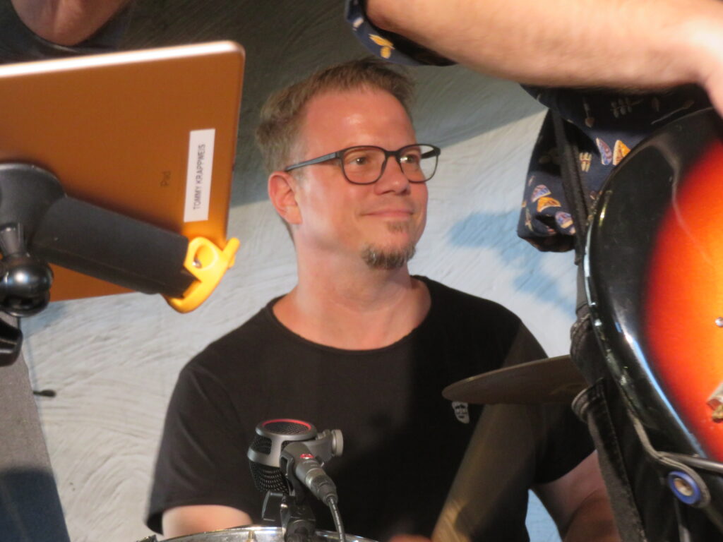 Florian Oswald sitzt am Schlagzeug, lächelt und trägt ein schwarzes T-Shirt. Auf der Nase hat er eine schwarze Brille. Vor ihm erkennt man einen Ständer mit einem iPad sowie den Unterarm und ein Stück der E-Gitarre von Muxi.