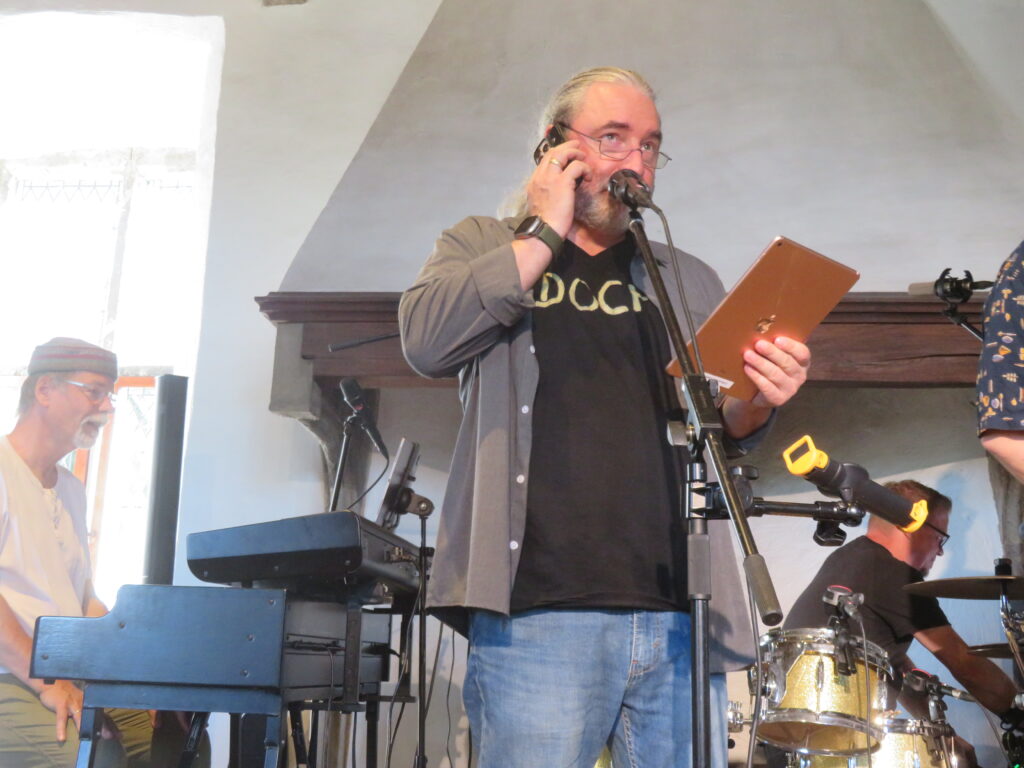 Tommy steht mit iPad und Handy am Ohr vor dem Microphon, die Lesebrille auf der Nasenspitze. Er trägt ein graues Hemd über dem schwarzen DOCH.-Shirt und Jeans. Im Hintergrund warten Lenzi amKeyboard und Flo am Schlagzeug auf ihren weiteren Einsatz. Dahinter ist die mittelalterliche Dunstabzugshaube zu sehen.