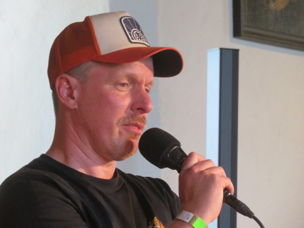 Norman schaut leicht nach unten und spricht gerade in ein Microphon. Er hat das rot-beige Cappy auf und trägt ein schwarzes T-Shirt. Hinter ihm sieht man die weiße Burgwand und einen dunklen Pfosten.