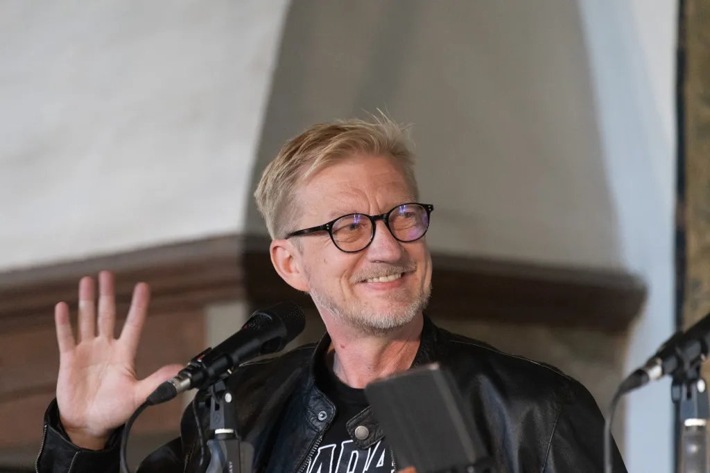 Detlef winkt jemandem zu und schaut rechts an der Kamera vorbei. Man sieht seine Zähne bei dem Versuch zu lächeln. Er trägt ein Dada-Welf-Shirt und eine schwarze Lederjacke. Im Hintergrund ist eine mittelalterliche Dunstabzugshaube zu sehen.
