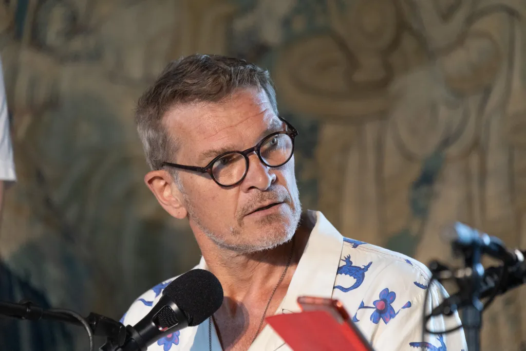 Götz schaut nachdenklich zur Seite durch seine schwarze Brille. Er hat ein weißes Hemd mit blauen Blumen und Salamandern an. Vor ihm stehen zwei Microphone und das rote iPad von Kai ist zu sehen. Im Hintergrund hängt ein mittelalterlicher Wandteppich.