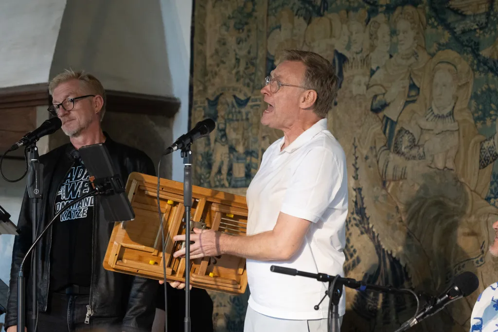 Thomas steht mit einem Holzklappbeistelltischlein vor dem Microphon, den Mund weit geöffnet. Er trägt ein weißes Poloshirt und graue Hosen. Im Hintergrund sieht man Detlef mit seinem Dada-Welf-Shirt und der Lederjacke, alles schwarz, und den mittelalterlichen Wandteppich.